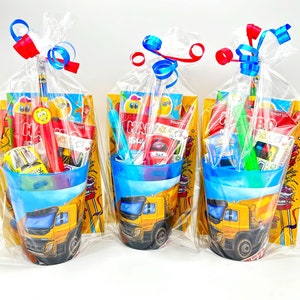 May include: Three clear plastic bags filled with party favours for a construction themed birthday party. Each bag contains a small blue cup with a yellow dumper truck design, a small colouring book, a pencil, and other small toys. The bags are tied with blue and red ribbon.