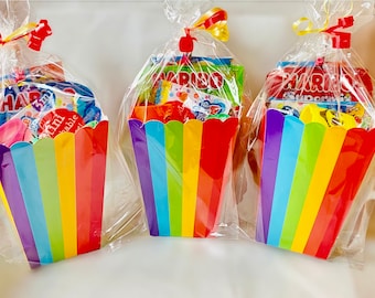 Może przedstawiać: Trzy pudełka na popcorn w paski tęczy wypełnione małymi zabawkami i słodyczami. Pudełka są owinięte w przezroczysty celofan i związane żółtą i czerwoną wstążką.