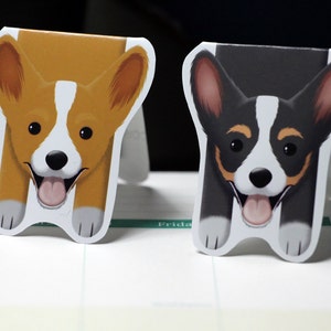 Puede incluir: Dos adorables marcadores de perro de dibujos animados. Un marcador presenta un corgi marr&oacute;n y blanco con la lengua rosa que sobresale. El otro marcador presenta un corgi negro y blanco con la lengua rosa que sobresale.