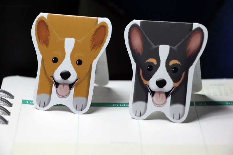 Puede incluir: Dos adorables marcadores de perro de dibujos animados. Un marcador presenta un corgi marr&oacute;n y blanco con la lengua rosa que sobresale. El otro marcador presenta un corgi negro y blanco con la lengua rosa que sobresale.