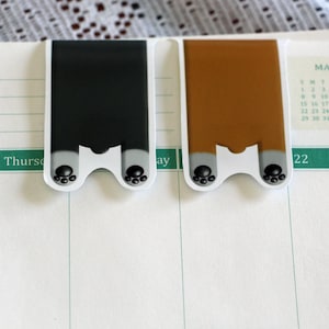 Puede incluir: Dos marcadores de p&aacute;gina con forma de pata, uno negro y otro marr&oacute;n, con huellas de patas negras en la parte inferior. Los marcadores est&aacute;n colocados en una p&aacute;gina blanca de un calendario con el mes de marzo visible.
