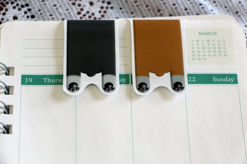 Puede incluir: Dos marcadores de p&aacute;gina con forma de pata, uno negro y otro marr&oacute;n, con huellas de patas negras en la parte inferior. Los marcadores est&aacute;n colocados en una p&aacute;gina blanca de un calendario con el mes de marzo visible.