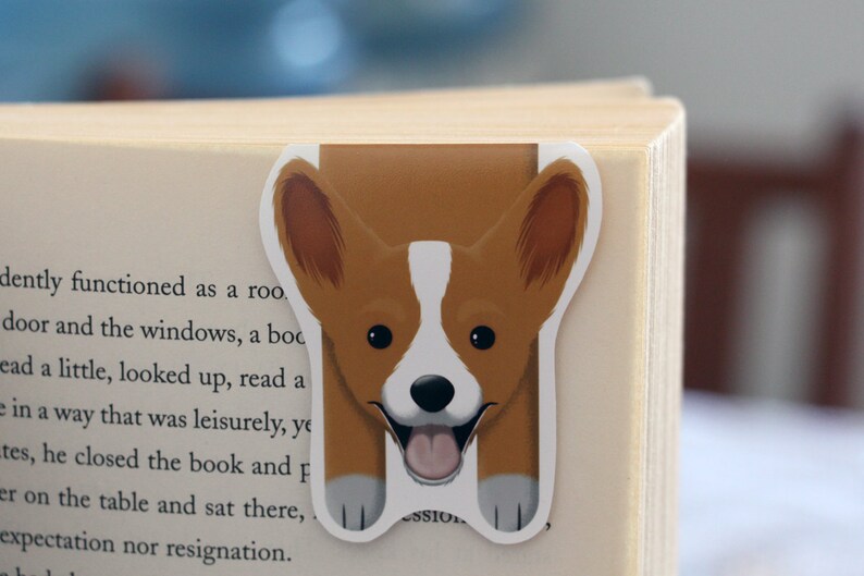 Puede incluir: Un marcap&aacute;ginas de corgi marr&oacute;n y blanco con una expresi&oacute;n feliz. El marcap&aacute;ginas tiene forma de corgi con la cabeza sobresaliendo de la parte superior del libro.