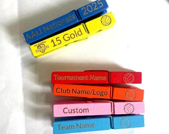 Puede incluir: Una pila de coloridas pinzas de madera. Las pinzas están pintadas en varios colores, incluyendo azul, amarillo, rojo, naranja, rosa y turquesa. Cada pinza tiene texto y un gráfico de voleibol. El texto incluye "AAU Nationals 2025", "15 Gold", "Tournament Name", "Club Name/Logo", "Custom" y "Team Name".