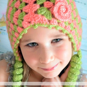 May include: A young girl wearing a pink and green crocheted hat with a flower design. The hat has long crocheted tassels.