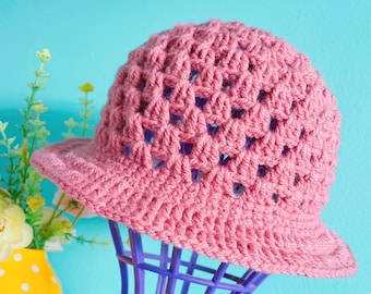 May include: A pink crocheted sun hat with a wide brim and an open weave pattern. The hat is displayed on a purple stand.