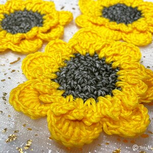Op de afbeelding: Drie gehaakte zonnebloemversieringen met gele bloemblaadjes en grijze centra. De zonnebloemen bevinden zich op een witte achtergrond met gouden glitter.