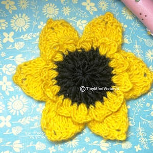 Op de afbeelding: Een gehaakte zonnebloem met gele bloemblaadjes en een zwart midden. De bloem is op een blauwe achtergrond met een wit bloemenpatroon.