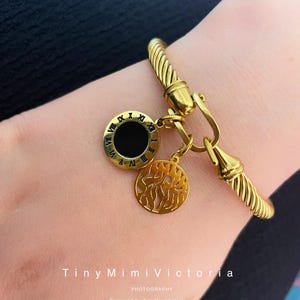 May include: Gold-toned bracelet with a twisted rope design. The bracelet features two dangling charms: one with Roman numerals and a black center, and another with a decorative cutout. The bracelet is secured with a hook clasp.
