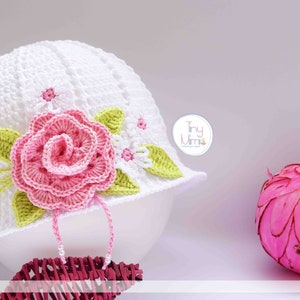 May include: White crocheted sun hat with a pink rose and green leaves. The hat has a white crocheted brim and a pink ribbon tied around the crown. The hat is decorated with a pink rose and green leaves. The hat is sitting on a brown wicker surface. The text "Tiny Mimi" is visible in the image.