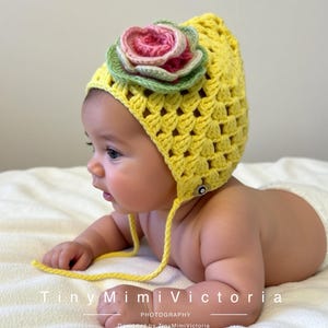 Pode incluir: Um gorro de bebê de crochê amarelo com uma grande flor rosa e verde. O gorro tem um padrão texturizado e laços sob o queixo. O bebê está deitado em um cobertor branco. O texto na imagem diz "Tiny Mimi Victoria Photography".