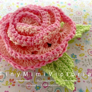 Peut inclure: Une rose rose en crochet avec des feuilles vertes. La rose est faite avec un fil rose clair et un fil rose foncé. Les feuilles sont faites avec un fil vert clair. La rose est sur un fond blanc avec un motif floral. Le texte "Tiny Mimi Victoria" est écrit sous la rose.