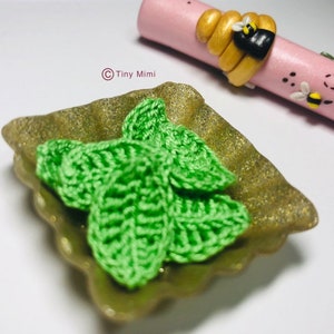 May include: A small, gold-colored dish with a scalloped edge holds three green crocheted leaves. The dish is on a white surface.