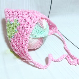 Puede incluir: Un gorro de bebé de ganchillo rosa con un diseño de corazón verde y una bola de lana rosa a juego.