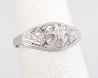 Pode incluir: Um anel de prata com um diamante central e detalhes gravados. O anel apresenta um design floral em torno do diamante principal, com diamantes menores e padrões geométricos na banda. O anel está sobre um fundo branco.