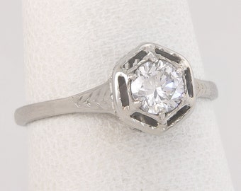 May include: A silver ring with a hexagonal setting and a round, clear gemstone. The band features etched leaf designs. The ring is displayed on a white background.