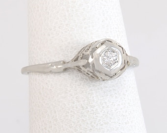 May include: A delicate, white metal ring with a filigree design. The ring features a hexagonal setting with a small, clear gemstone at its center. The band is thin and tapers towards the back.