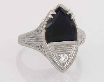 May include: A vintage-style ring with an elongated, shield-shaped black gemstone. The ring features intricate filigree detailing on the band and around the main stone. A small, round diamond is set at the bottom of the black stone.