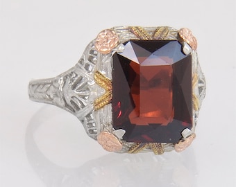 May include: A vintage-style ring featuring a large, rectangular, dark red gemstone. The ring has a silver-colored filigree band with gold and rose gold accents around the gemstone, creating an antique aesthetic. The gemstone is the focal point.