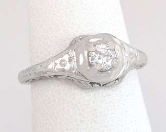 May include: A silver engagement ring with a round, clear gemstone. The ring features intricate filigree and engraved details on the band and setting. The ring is displayed on a white background.