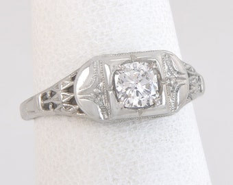 May include: A silver-toned ring with a square, clear gemstone at the center. The ring features intricate filigree and geometric designs on the band and setting. The ring is displayed on a white background.