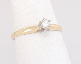 May include: A gold engagement ring featuring a single, clear diamond. The ring has a simple band and a four-prong setting. The ring is displayed against a white backdrop. The ring is likely made of gold.