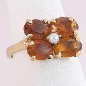 May include: A gold ring featuring a cluster of four oval-cut, amber-colored gemstones arranged around a central pearl. The ring has a simple, elegant band and a floral-like design.