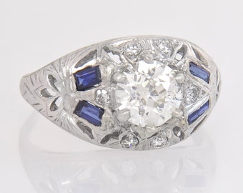 May include: A vintage-style ring with a large, round, clear diamond as the centerpiece. The ring is set in a silver-colored metal and features smaller diamonds and rectangular blue gemstones. The band has an engraved floral design.