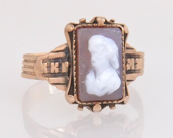 May include: A gold-toned ring featuring a rectangular cameo with a white profile carving set within a decorative frame. The ring's band has detailed engravings, and the cameo has a light brown background.