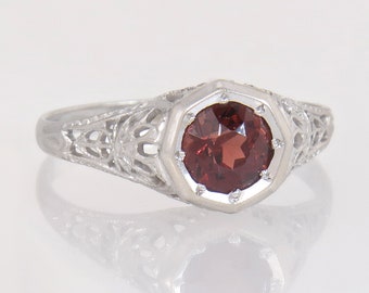 May include: A silver ring with a round, deep red gemstone set in an octagonal bezel. The band features intricate filigree detailing. The ring is displayed on a reflective surface, highlighting its design and color.