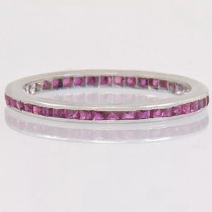 May include: A silver ring with a continuous band of square-cut, vibrant pink gemstones. The ring's polished metal reflects the light, highlighting the gemstones' color and the ring's smooth, rounded edges. A classic piece of jewelry.