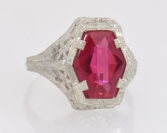 May include: A silver ring with a large, faceted, ruby-red gemstone. The ring has an ornate, filigree design around the gemstone and on the band. The gemstone is a hexagonal shape.