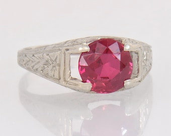 May include: A vintage-style ring with a large, faceted, round, ruby-red gemstone. The ring's band is white metal, possibly silver or platinum, with intricate engraved floral and geometric patterns. The gemstone is the focal point.