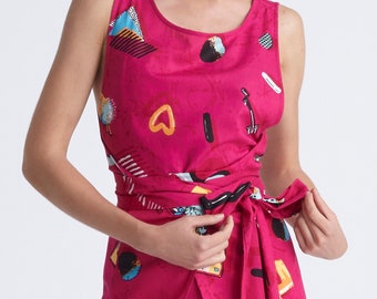 May include: A sleeveless fuchsia dress with a wrap-around waist. The dress features a repeating pattern of colorful abstract shapes, including hearts, circles, and geometric designs. The dress is made of a lightweight fabric.