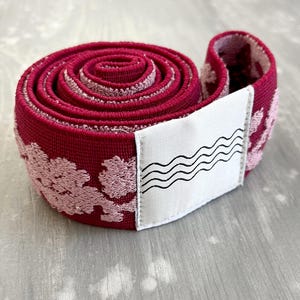 May include: A coiled roll of deep red elastic fabric featuring a textured, light pink floral pattern throughout. A white rectangular fabric label is attached to the outer edge, displaying a black graphic print of four horizontal wavy lines.