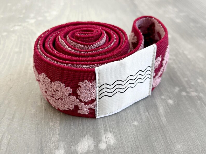 May include: A coiled roll of deep red elastic fabric featuring a textured, light pink floral pattern throughout. A white rectangular fabric label is attached to the outer edge, displaying a black graphic print of four horizontal wavy lines.