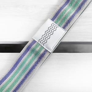 May include: A fabric strap featuring a repeating pattern of light green and purple stripes on a textured beige background. A white rectangular patch is centered on the strap, displaying a black graphic design of four parallel wavy lines.