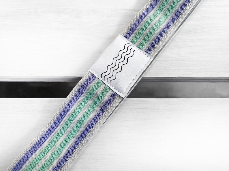 May include: A fabric strap featuring a repeating pattern of light green and purple stripes on a textured beige background. A white rectangular patch is centered on the strap, displaying a black graphic design of four parallel wavy lines.