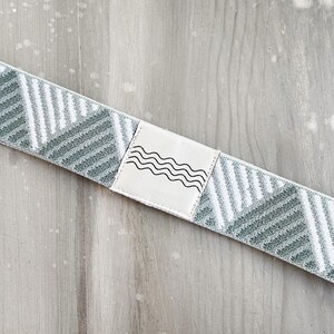 May include: A stretchable fabric band featuring a repeating geometric pattern of sage green and white diagonal stripes. A rectangular white patch is stitched onto the center, displaying a black line drawing of four horizontal wavy lines.