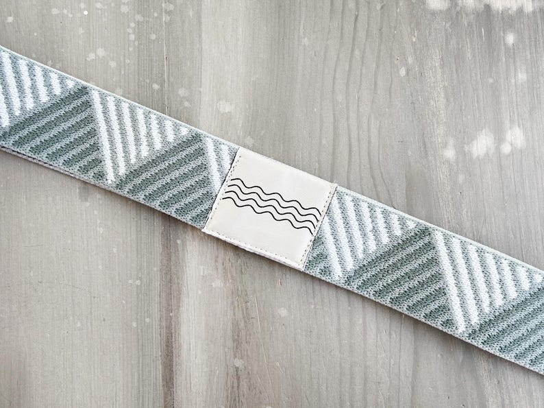 May include: A stretchable fabric band featuring a repeating geometric pattern of sage green and white diagonal stripes. A rectangular white patch is stitched onto the center, displaying a black line drawing of four horizontal wavy lines.