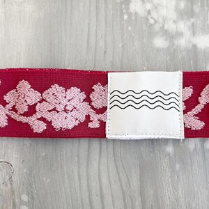 May include: A deep red elastic band featuring a repeating light pink floral embroidery pattern. A white square fabric tag is stitched onto the center of the band, displaying a black graphic print of four horizontal, parallel wavy lines.