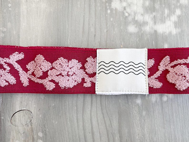 May include: A deep red elastic band featuring a repeating light pink floral embroidery pattern. A white square fabric tag is stitched onto the center of the band, displaying a black graphic print of four horizontal, parallel wavy lines.