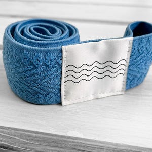 May include: A coiled blue elastic band featuring a textured floral pattern and a white rectangular fabric patch sewn onto the front. The patch displays a black graphic print consisting of four horizontal wavy lines stacked vertically.