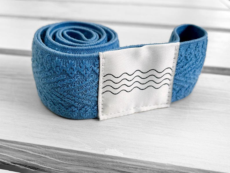 May include: A coiled blue elastic band featuring a textured floral pattern and a white rectangular fabric patch sewn onto the front. The patch displays a black graphic print consisting of four horizontal wavy lines stacked vertically.