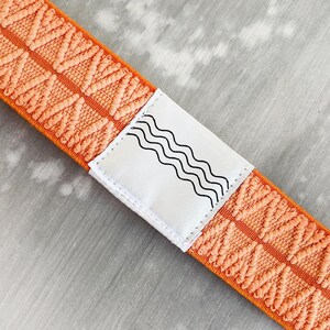 May include: An orange elastic strap featuring a repeating textured chevron pattern. A white rectangular fabric label is stitched onto the center of the strap, displaying a black graphic design consisting of four parallel wavy horizontal lines.