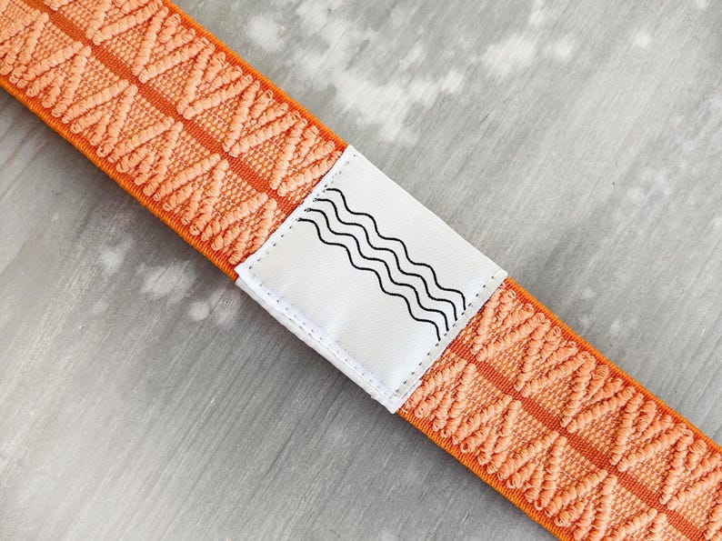 May include: An orange elastic strap featuring a repeating textured chevron pattern. A white rectangular fabric label is stitched onto the center of the strap, displaying a black graphic design consisting of four parallel wavy horizontal lines.