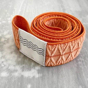 May include: A coiled orange elastic resistance band featuring a textured diamond-shaped weave pattern. A white rectangular fabric label is attached to the end of the band, displaying a black graphic print of four horizontal wavy lines.