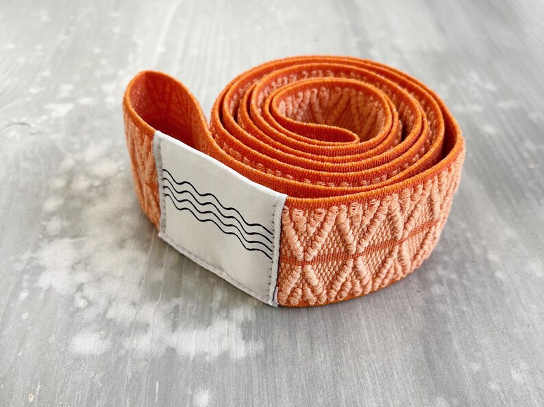 May include: A coiled orange elastic resistance band featuring a textured diamond-shaped weave pattern. A white rectangular fabric label is attached to the end of the band, displaying a black graphic print of four horizontal wavy lines.