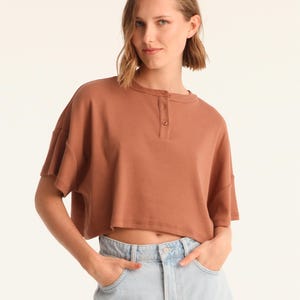 May include: A brown, cropped t-shirt with short sleeves and a henley collar. The top is paired with light blue denim shorts. The model is standing in front of a white background. The shirt has a relaxed fit.
