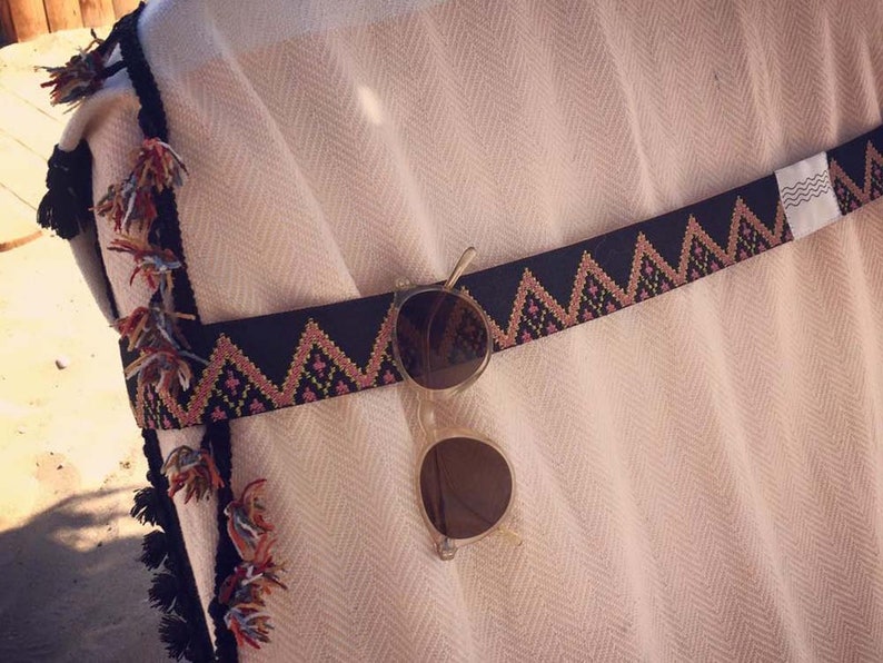 May include: A cream-colored textile with a herringbone pattern features a black woven strap with a pink and orange zigzag design. The strap is adorned with multicolored tassels along the edge and holds a pair of round, light-colored sunglasses.